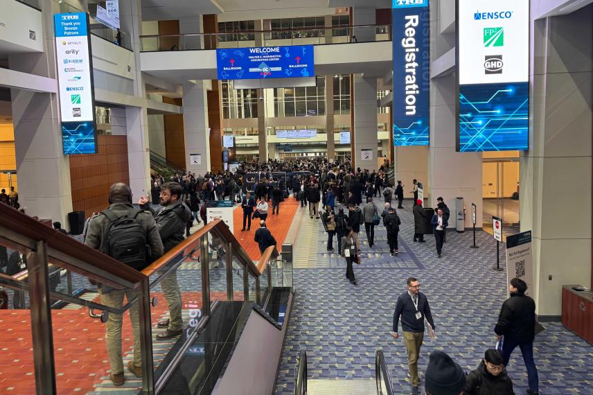 Large convention center atrium with crowds of attendees, blue registration signage, and corporate sponsor logos displayed on walls and digital screens.
