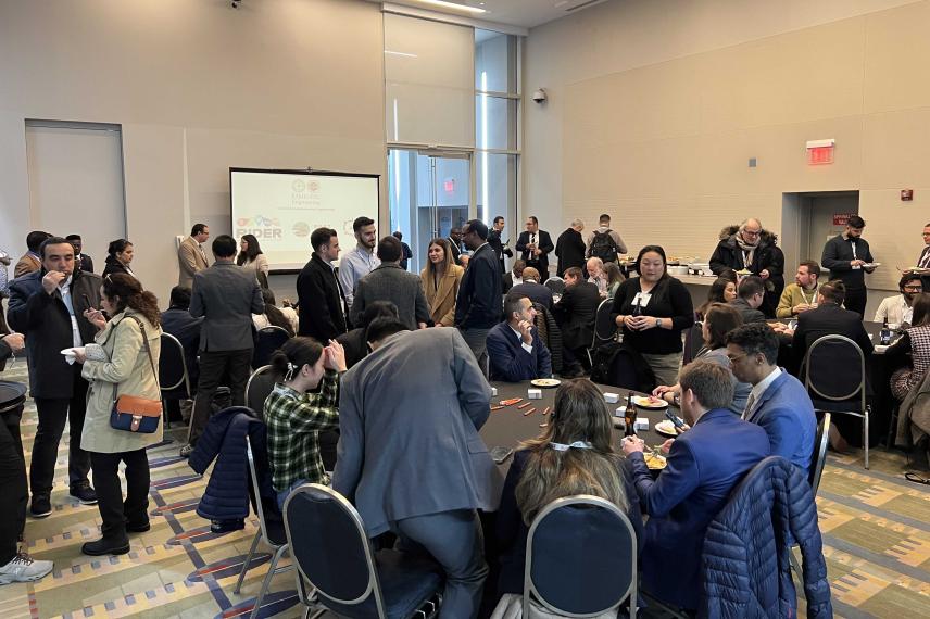 A crowded conference or networking event in a modern venue with attendees seated at tables and standing, with a presentation screen visible in the background.