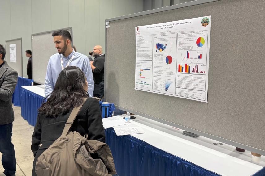 A presenter in a light blue shirt explains a research poster on workforce skills in transportation to a seated visitor at an academic conference.