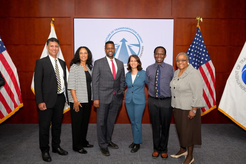 Six professionals stand in front of a "Future of Transportation Summit" banner, flanked by American flags and state flags in a formal setting.