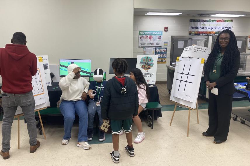 A woman facilitates an educational program with students in a classroom, displaying a tic-tac-toe game board while students engage with interactive content on a nearby screen.
