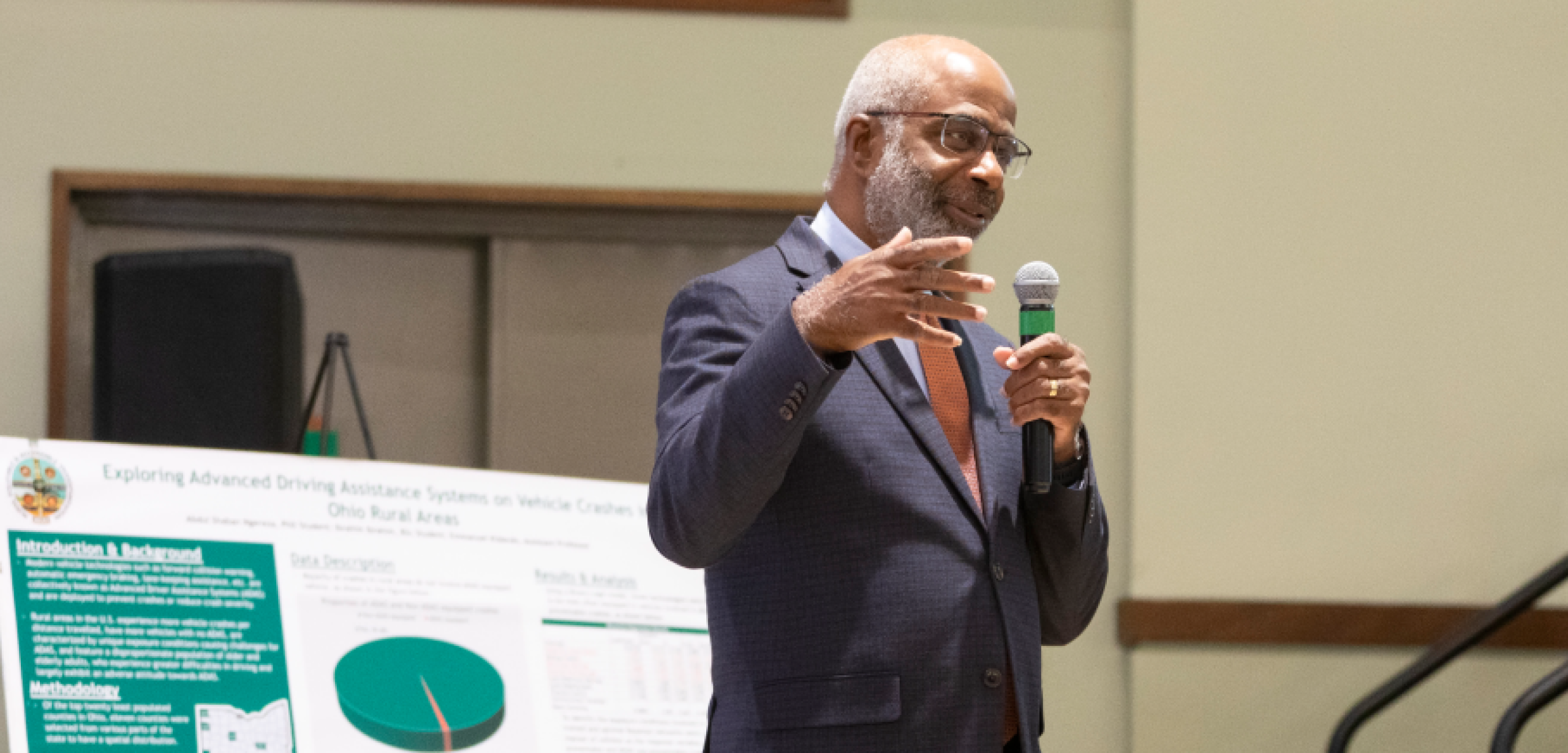 A man is speaking at the front of a rom holding a microphone. There are partial views of posters with graphs and information behind him.