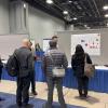 Visitors examine a research poster at an academic conference or poster session in a convention center hall with blue-draped tables.