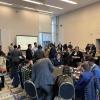 A crowded conference or networking event in a modern venue with attendees seated at tables and standing, with a presentation screen visible in the background.