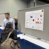 A presenter in a light blue shirt explains a research poster on workforce skills in transportation to a seated visitor at an academic conference.