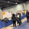 Attendees view research posters at an academic conference in a large hall with blue-draped tables and displayed presentation boards.