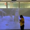 Person in yellow shirt standing before large projection display showing an urban underpass scene in blue tones within a modern glass-walled room.